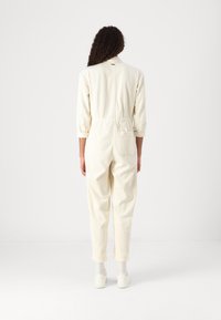 Mono de pana color crema con cuello tipo camisa, mangas largas y cintura elástica; cuenta con dos bolsillos traseros y piernas ajustadas.