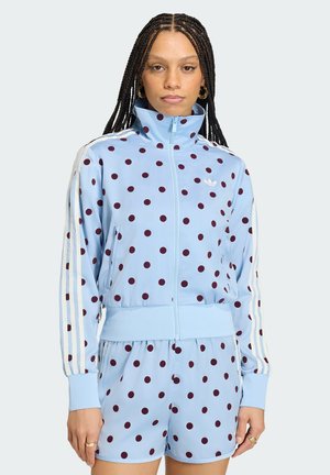 Donna con lunghe trecce che indossa una giacca sportiva azzurra a pois con strisce bianche e pantaloncini abbinati, in piedi davanti a uno sfondo semplice.