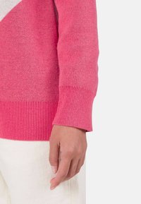 Różowy sweter z dzianiny o gradientowym wzorze i prążkowanymi mankietami, zestawiony z białymi materiałowymi spodniami. Tekstura wydaje się miękka i jednolita.