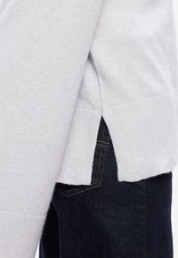 Ανοιχτό γκρι πουλόβερ με ριμπ μανσέτες και πλαϊνές σχισμές, συνδυασμένο με σκούρες τζιν παντελόνες. Μαλακή υφή ορατή στο ύφασμα.