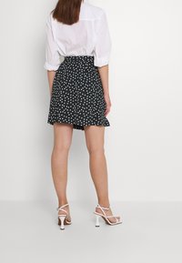 Jupe patineuse noire à pois blancs, en tissu fluide, avec un détail de ceinture, associée à des sandales à talons hauts blanches à lanières, vue de dos.