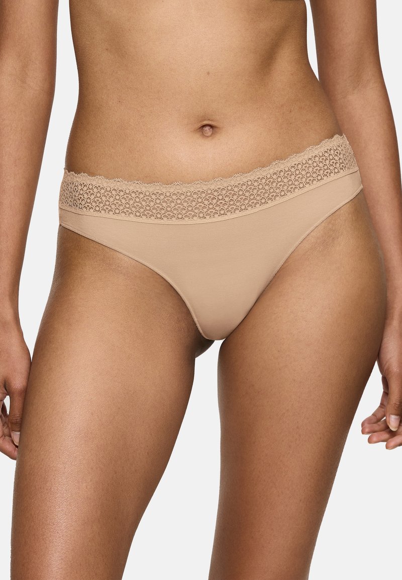 Femme portant un sous-vêtement bikini beige orné de dentelle, montrant le ventre et le haut des cuisses sur un fond neutre.