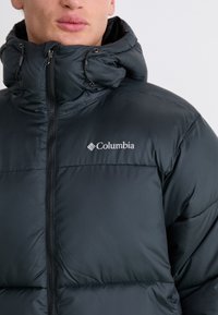 Schwarze Steppjacke mit hohem Kragen und Reißverschluss, die ein Logo auf der Brust hat. Der Stoff wirkt glatt und hat einen leichten Glanz.