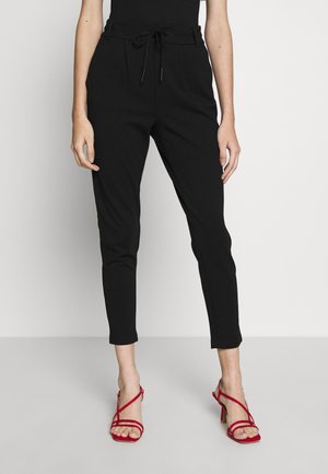 Donna che indossa pantaloni neri aderenti con coulisse in vita e sandali rossi con tacco alto e cinturini, in piedi su un pavimento grigio chiaro.