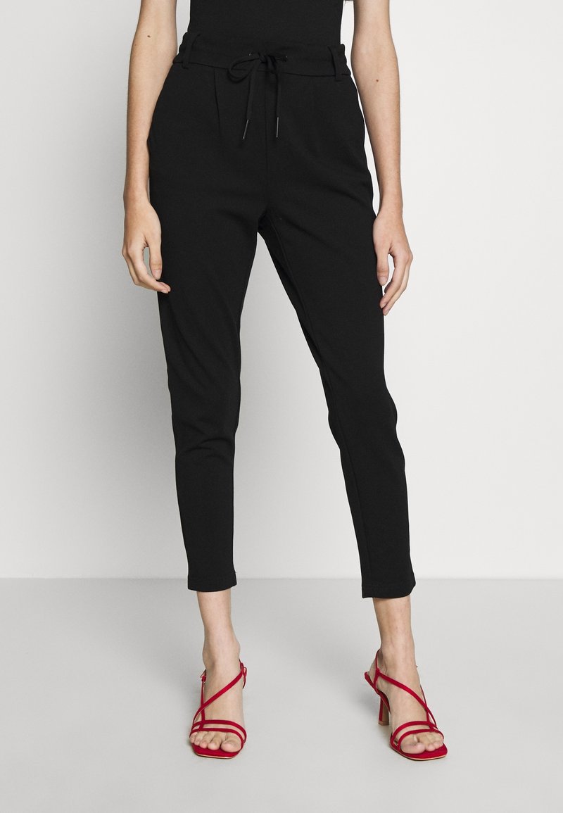 Femme portant un pantalon noir ajusté avec une taille à cordon et des sandales rouges à talons hauts à lanières, debout sur un sol gris clair.