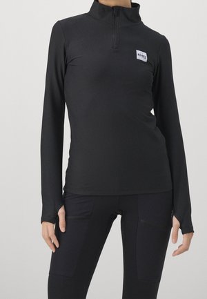 Personne portant un haut noir ajusté à manches longues avec des trous pour les pouces et un legging noir avec poches, debout devant un fond clair uni.