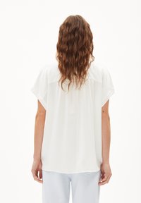 White, short-sleeve top with a relaxed fit, featuring a rounded hem and smooth fabric, shown from the back with wavy hair visible.