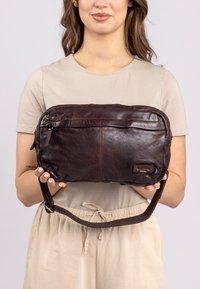 Brown leather crossbody bag with a zip closure, textured surface, and adjustable fabric strap. Features a front pocket and logo detail.