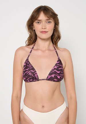 Kobieta w różowym i ciemnym wzorzystym topie bikini z zakładką na szyję oraz gładkich, białych, wysoko wyciętych majtkach od bikini na jednolitym tle.