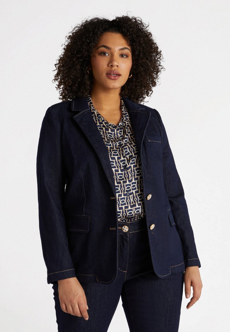 Blazer en denim foncé avec un design ajusté, revers crantés, accents de boutons dorés et deux poches avant. Assorti à un chemisier à motifs en dessous.