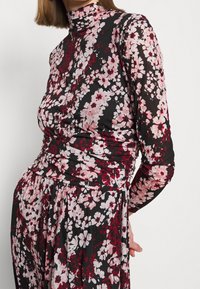 Pitkähiha, korkeakauluksinen paita mustasta kankaasta, jota koristaa vaaleanpunaisista ja punaisista kukista koostuva kukkakuosi. Vyötäröltä ker gathered yksityiskohdat.