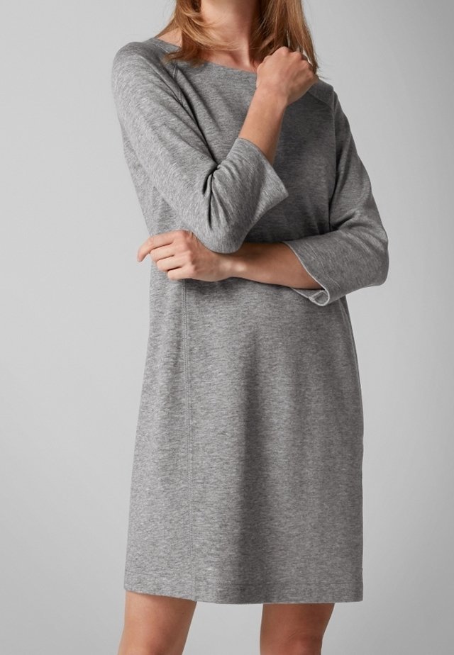 Femme portant une robe grise ample à manches longues, debout avec les bras croisés devant un fond gris uni.