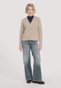 Blonde Frau trägt eine beige, geknöpfte Strickjacke über einem schwarzen Oberteil, weit geschnittene, hellblaue Jeans und braune Schuhe und steht vor einem hellen Hintergrund.