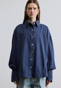 Μπλε κλασικό τζιν μπουφάν σε μεγάλη διάσταση, με κουμπιά στο εμπρός μέρος, μεγάλο γιακά και μακριά μανίκια. Ασημένια κουμπιά και διακριτικές ραφές.