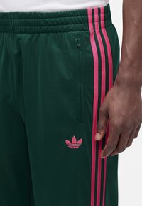 Σκούρο πράσινο παντελόνι Adidas με ροζ ρίγες και λογότυπο, ελαστική μέση, πλαϊνή τσέπη με φερμουάρ και το δεξί χέρι ενός ατόμου που ακουμπά δίπλα στο παντελόνι.