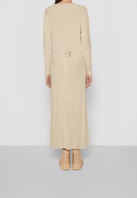 Robe longue en maille beige avec manches longues et ceinture à la taille. Texture lisse, coupe ample et fente latérale en bas. Associée à des bottines beige.