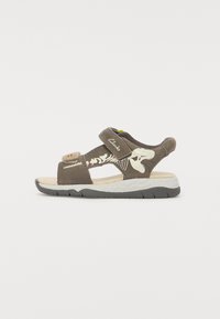 Bruna mockaskinnsandaler med ett kardborreband, prydda med ett dinosaurieskelettdesign på sidan och en vadderad vit sula med svarta detaljer.