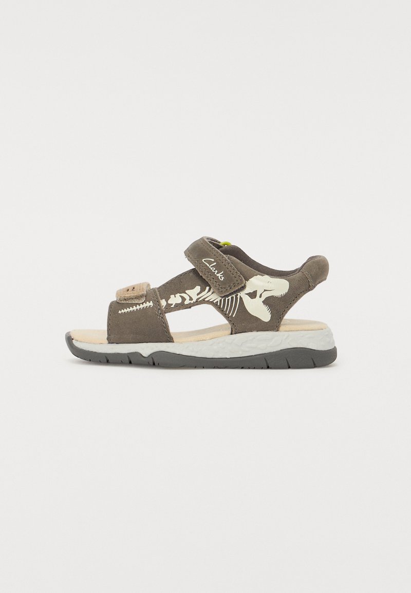Bruna mockaskinnsandaler med ett kardborreband, prydda med ett dinosaurieskelettdesign på sidan och en vadderad vit sula med svarta detaljer.