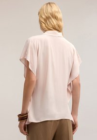 Blusa corta a maniche corte di un rosa pallido, con una vestibilità morbida e un tessuto drappeggiato. Presenta un colletto e un orlo curvo.