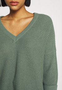 Maglione verde lavorato a maglia con scollo a V, caratterizzato da una superficie testurizzata e maniche ampie. Il tessuto appare morbido e traspirante.