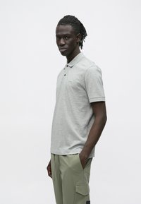 Młody mężczyzna z dredami, ubrany w jasnoszarą koszulkę polo i oliwkowe spodnie bojówki, stoi z jedną ręką w kieszeni.