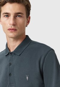 Mørkegrøn polo shirt lavet af tekstureret stof, med en krave, tre knapper og et lille broderet kraniumlogo på venstre bryst.