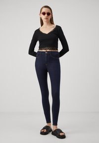 Top a maniche lunghe nera e cropped con bordi in pizzo, abbinato a jeans skinny blu scuro. Il modello indossa sandali neri e occhiali da sole rosa.