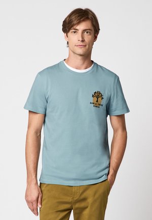 Junger Mann trägt ein hellblaues T-Shirt mit Sonnen- und Baum-Motiv, kombiniert mit senffarbenen Hosen, vor einem grauen Hintergrund stehend.