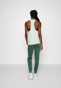 Ljusgrön racerback linne kombinerat med mörkgröna träningsbyxor med vita sidostråkar; modellen visas bakifrån, iklädd vita sneakers.