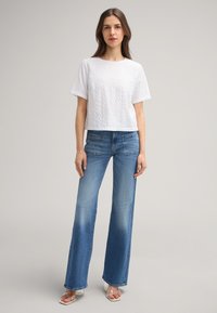 Witte cropped t-shirt met textuurpatroon, gecombineerd met lichtblauwe flare jeans met achterzakken. Model draagt doorzichtige sandalen.