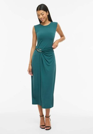 Vrouw draagt een mouwloze teal midi-jurk met geplooide taille en zwarte enkellaarzen met riempje, staand en glimlachend tegen een witte achtergrond.