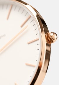 Różowe złote zegarek z białą tarczą, z wydłużonymi znacznikami godzin, cienkimi wskazówkami i fakturowaną koronką z boku. Minimalistyczny design.