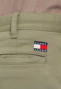 Κλείσιμο της μέσης χακί παντελονιού με θηλιά ζώνης και πίσω τσέπη, με ετικέτα λογότυπου Tommy Jeans σε ναυτικό μπλε, άσπρο και κόκκινο.