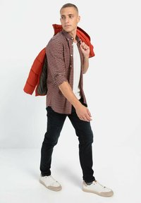 Geruit overhemd in rood, zwart en wit, gecombineerd met een wit T-shirt, donkere spijkerbroek en witte sneakers met beige accenten, en een oranje pufferjack.
