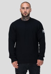 Zwarte geribbelde gebreide trui met lange mouwen, ronde hals en logopatch op de linkerarm. Gemaakt in combinatie met verouderde donkerdenimjeans.