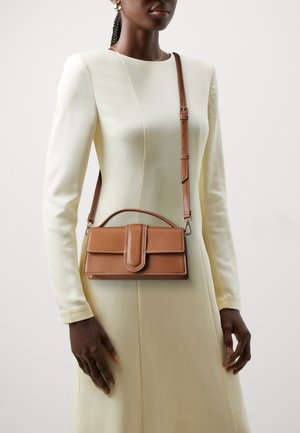 Vrouw draagt een crèmekleurige jurk met lange mouwen en een gestructureerde, bruine lederen crossbodytas met een handvat bovenop en een gespdetail.