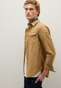 Lichtbruine katoenen blouse met een knoopsluiting aan de voorkant, twee borstzakken en een licht ontspannen pasvorm. Draag het over een wit T-shirt en blauwe denim jeans.