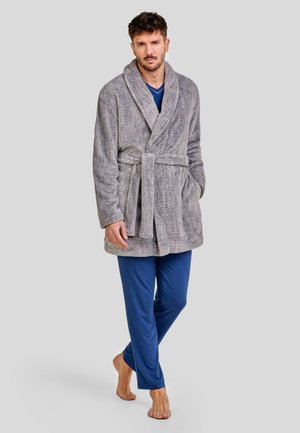 Bata de color gris con patrón de espiga, con cuello chal, cintura con cinturón y bolsillos laterales. Combinada con pantalones de pijama azules y descalza.