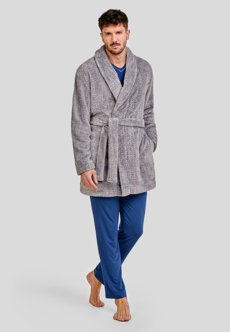 Bata de color gris con patrón de espiga, con cuello chal, cintura con cinturón y bolsillos laterales. Combinada con pantalones de pijama azules y descalza.