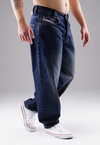Man wearing loose dark blue jeans and white sneakers, standing against a plain light gray background.