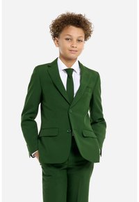 Completo verde con una vestibilità su misura, giacca monopetto, rever a taglio e pantaloni abbinati. Camicia bianca e cravatta scura completano il look.