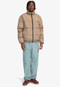 Tan pufferjakke med høj krave og lynlås foran, med kviltede sektioner. Båret over lyseblå jeans og brune sko.