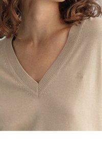 Beige V-hals trui met een gladde textuur, geribbelde halslijn en subtiele branding op de borst. Beschikt over fijne stiksels.