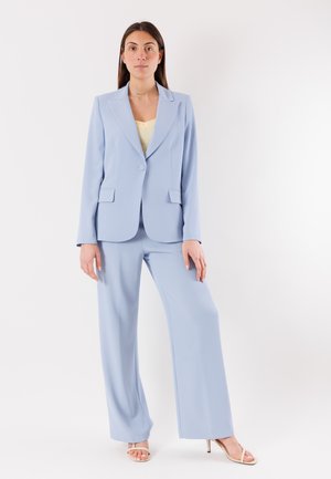 Donna in piedi che indossa un completo pantaloni azzurro con pantaloni a gamba larga, blazer con un solo bottone, top giallo e tacchi aperti beige su sfondo bianco.