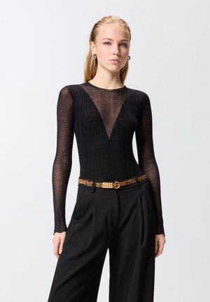 Tânără purtând un top negru transparent cu mâneci lungi și un decolteu adânc în V, pantaloni negri cu talie înaltă și o curea cu imprimeu leopard și cataramă aurie.