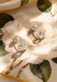 Silverblommformade örhängen med en klar ädelsten i mitten, presenterade på en blommönstrad tygbakgrund.