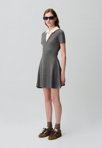 Robe grise avec une jupe évasée courte, des manches courtes et un col blanc. Associée à des chaussures brunes et des chaussettes rayées. Design minimaliste.