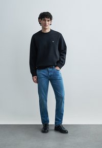 Svart sweatshirt med en liten vit logotyp, kombinerad med ljusblå jeans och svarta skor. Tyget verkar mjukt med en avslappnad passform.
