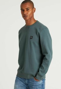 Felpa verde realizzata in materiale texturizzato, con scollo a girocollo e una piccola etichetta con logo nero sul lato sinistro del petto, abbinata a jeans blu.