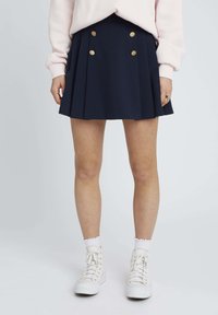 Jupe plissée bleu marine avec double boutons dorés à l'avant, associée à des baskets montantes blanches et des chaussettes côtelées.